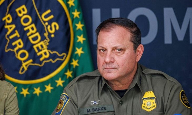 Border Patrol agent M. Banks in uniform at a press conference, with agency patches and a seals backdrop behind him.