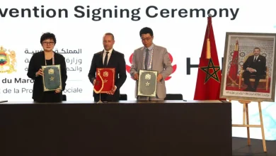 Three officials stand behind a table holding decorative folders during a signing ceremony, with the Moroccan flag nearby and a framed portrait on display.
