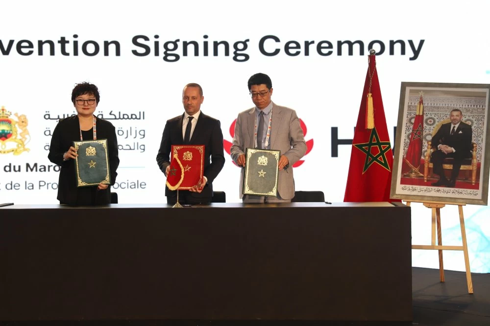 Three officials stand behind a table holding decorative folders during a signing ceremony, with the Moroccan flag nearby and a framed portrait on display.