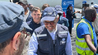 537971 | MCG24 | موقع إلكتروني مغربي شامل متجدد على مدار الساعة Man wearing a World Health Organization cap and vest smiles while surrounded by a crowd at a WHO UNHAS event near a plane.