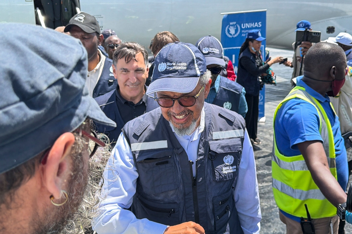 Man wearing a World Health Organization cap and vest smiles while surrounded by a crowd at a WHO UNHAS event near a plane.