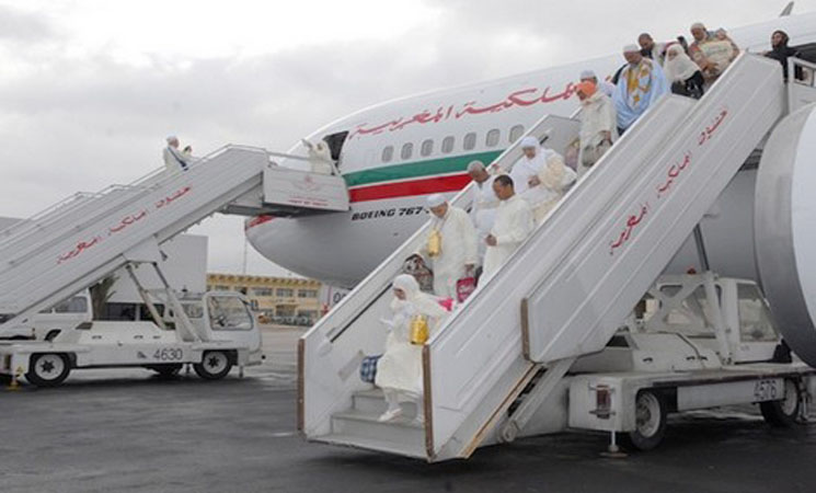 People in white traditional robes boarding a Boeing aircraft via mobile stairs at a tarmac; plane has a green-red-white stripe along the fuselage.