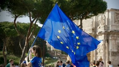 A person waves a large European Union flag at an outdoor gathering near a historic monument.
