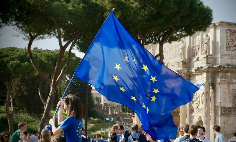 A person waves a large European Union flag at an outdoor gathering near a historic monument.