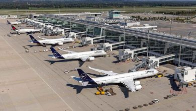 Aerial view of several Lufthansa airplanes parked at gates around a modern airport terminal with jet bridges and ground support equipment nearby.