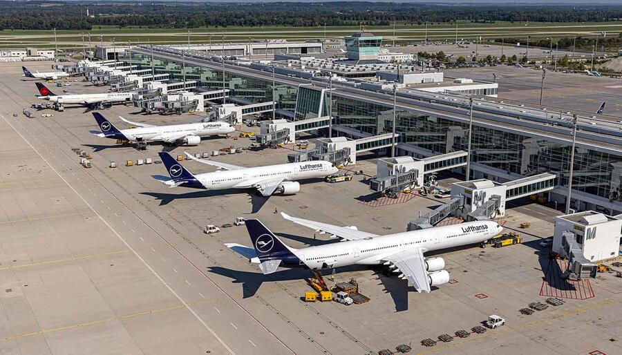 Aerial view of several Lufthansa airplanes parked at gates around a modern airport terminal with jet bridges and ground support equipment nearby.