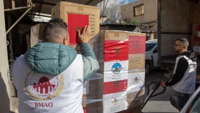 Two aid workers load a pallet of boxed supplies wrapped in plastic in a loading yard; one wears a white vest with the BMAQ logo.