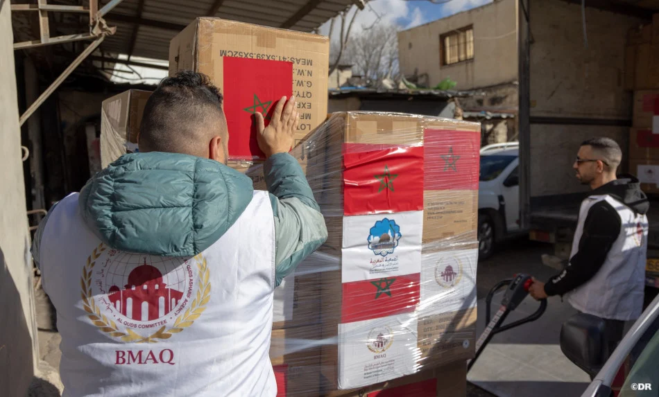 Two aid workers load a pallet of boxed supplies wrapped in plastic in a loading yard; one wears a white vest with the BMAQ logo.