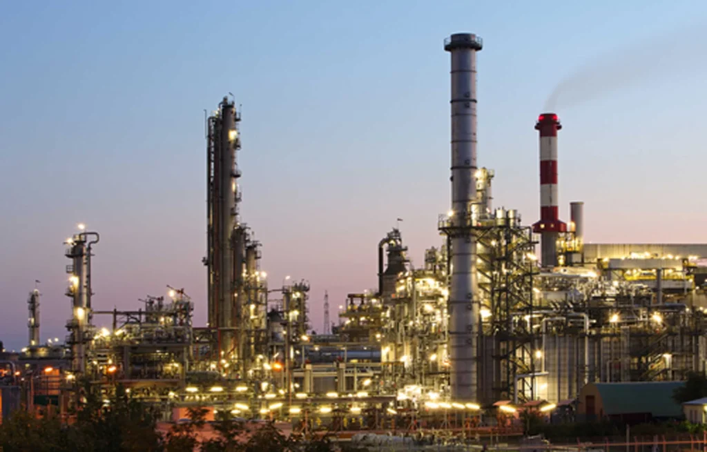 Industrial refinery complex with tall smokestacks and glowing lights at dusk.