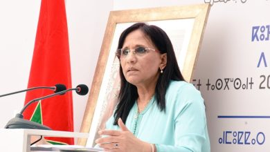 Woman in a light blue outfit speaking at a podium with two microphones, Moroccan flag in the background.