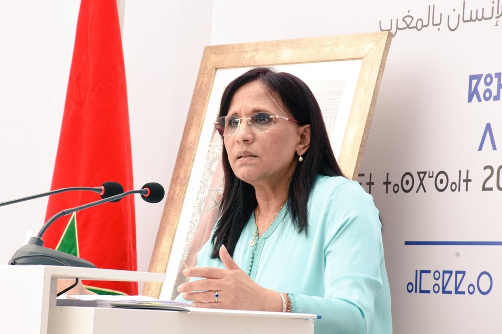 Woman in a light blue outfit speaking at a podium with two microphones, Moroccan flag in the background.