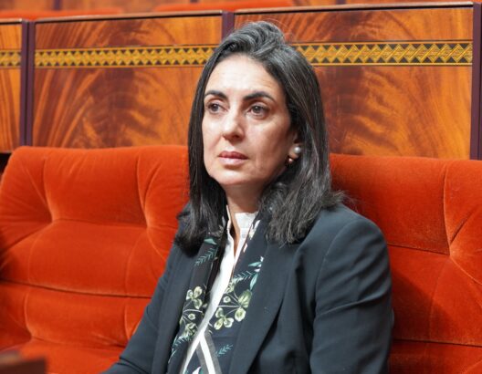 Middle-aged woman with dark hair in a navy blazer and floral scarf sits on a red-orange tufted couch, wood-paneled backdrop.