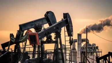 Oil field with large pumpjacks at sunset, silhouetted against an orange sky near processing equipment and fences.