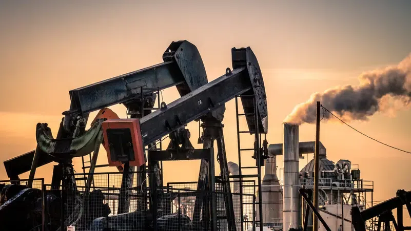 Oil field with large pumpjacks at sunset, silhouetted against an orange sky near processing equipment and fences.