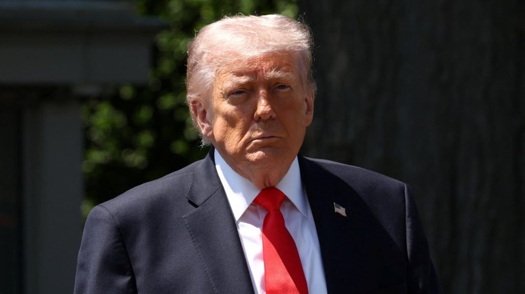 Man in a dark suit with a red tie and an American flag lapel pin, standing outdoors with a dark background.