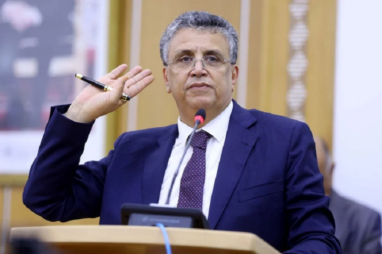 Man in a dark blue suit delivers a speech at a podium, raising a pen in his left hand with a microphone in front of him.