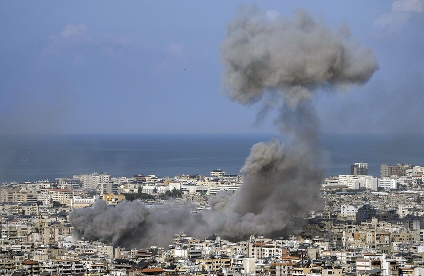 Large plume of smoke rising over a densely built coastal city, likely from an explosion near the shore.