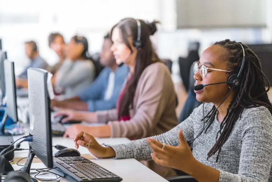 Customer service agent with headset at a computer in a busy call center environment.