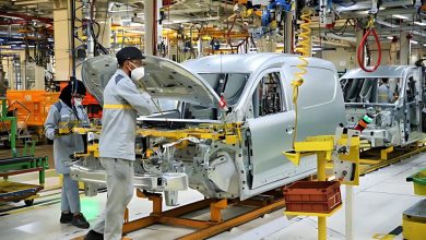 Automotive factory assembly line with silver car bodies on an orange conveyor; workers in masks inspect a vehicle under the hood.