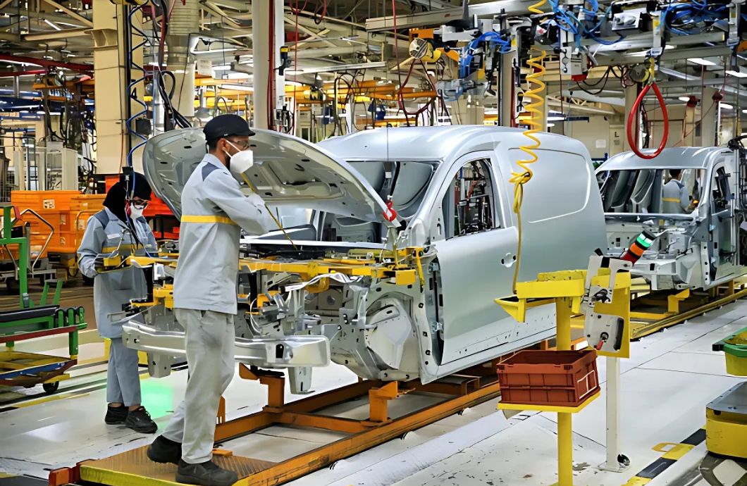 Automotive factory assembly line with silver car bodies on an orange conveyor; workers in masks inspect a vehicle under the hood.