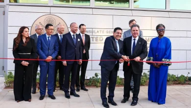 Dignitaries in suits at a ribbon-cutting outside a government building, two men cutting the red ribbon as others look on.