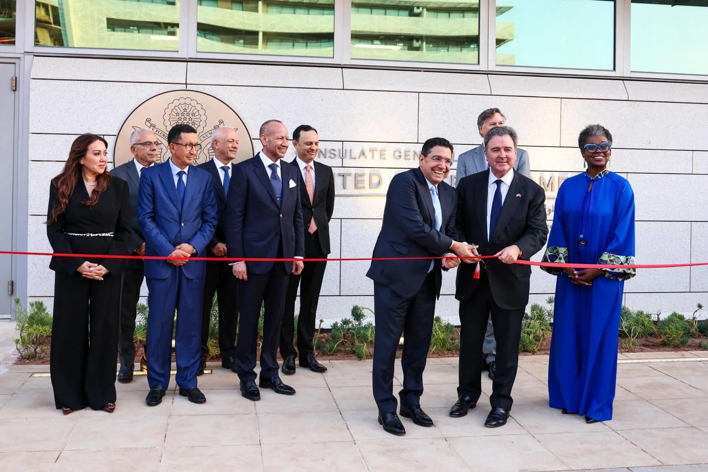 Dignitaries in suits at a ribbon-cutting outside a government building, two men cutting the red ribbon as others look on.