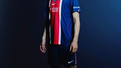 Full-body portrait of a male soccer player in a Paris Saint-Germain kit, standing against a dark blue gradient backdrop.