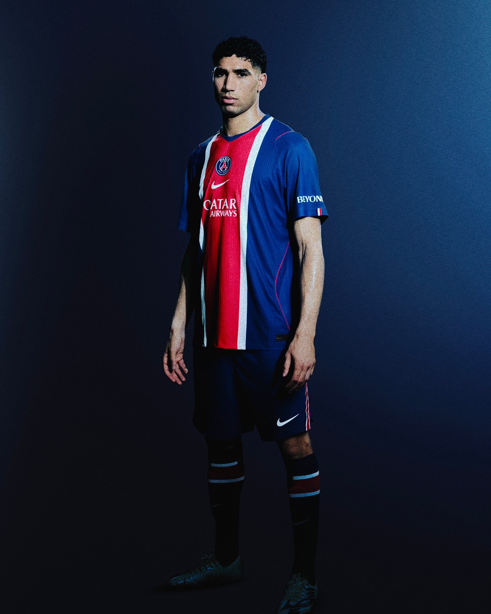 Full-body portrait of a male soccer player in a Paris Saint-Germain kit, standing against a dark blue gradient backdrop.