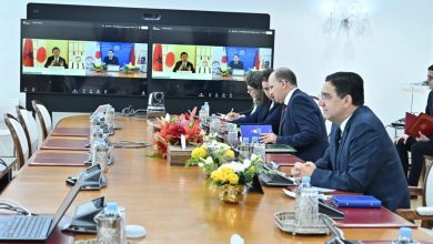 Formal meeting in a bright conference room with six professionals around a long table; two screens show a video conference with officials.
