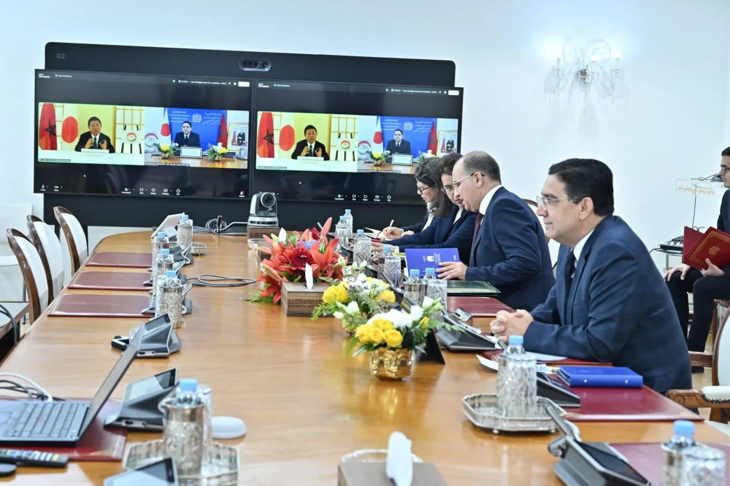 Formal meeting in a bright conference room with six professionals around a long table; two screens show a video conference with officials.