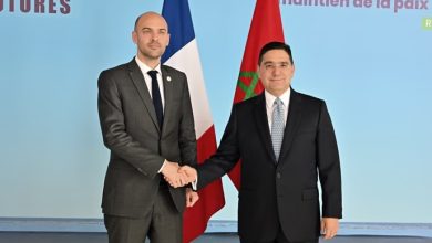 Two men in dark suits shake hands in front of French and Moroccan flags at a formal diplomatic event in a light-blue setting.