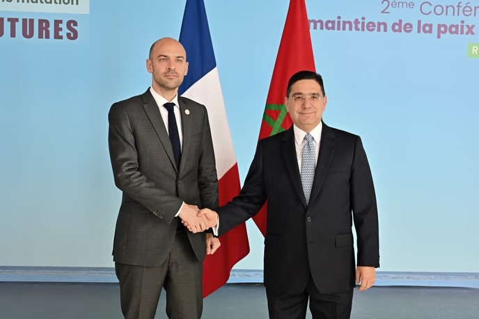 Two men in dark suits shake hands in front of French and Moroccan flags at a formal diplomatic event in a light-blue setting.
