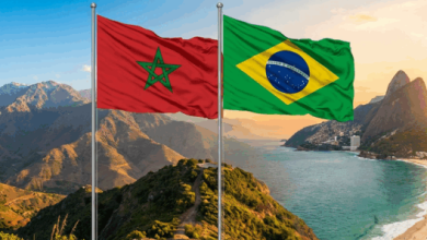 Moroccan and Brazilian flags waving on flagpoles above a coastal mountain landscape at sunset.