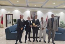 Five men in business suits pose in a formal lobby, presenting a wooden award plaque, with Moroccan flags and a framed portrait in the background.