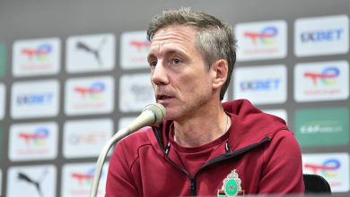 Man in a maroon jacket with a crest speaks into a microphone at a press conference, backdrop filled with sponsor logos.