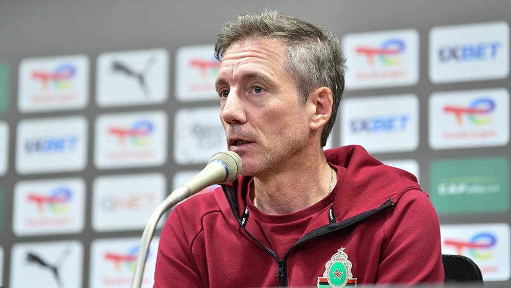 Man in a maroon jacket with a crest speaks into a microphone at a press conference, backdrop filled with sponsor logos.