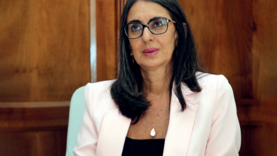 Woman with dark hair and glasses seated in a wood-paneled room, wearing a pale pink blazer and black top.