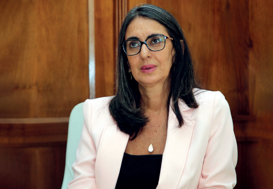 Woman with dark hair and glasses seated in a wood-paneled room, wearing a pale pink blazer and black top.