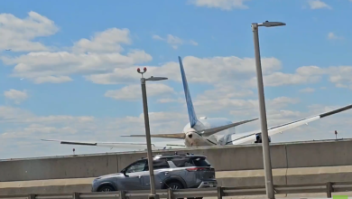 Airliner on a taxiway behind a low barrier, with a gray SUV driving past in the foreground under a blue sky.