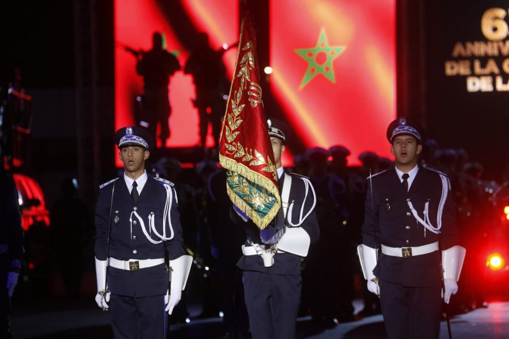 Three uniformed soldiers stand at attention during a nighttime parade, with a decorative red flag between them and a red-lit stage backdrop behind them.