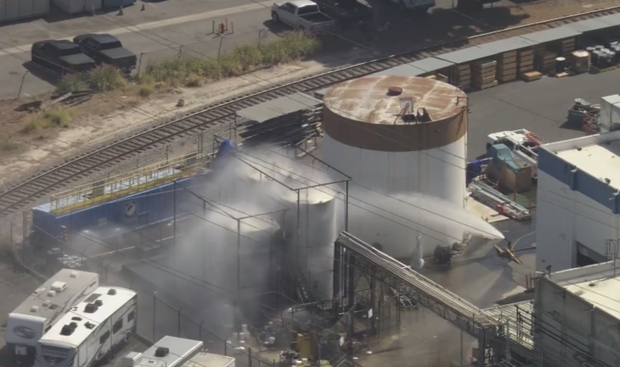 Industrial facility with large cylindrical storage tank and steaming vats near railroad tracks outside.