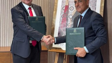 Two businessmen in suits shake hands after a formal agreement, each holding a green folder in a modern office setting.”] } {
