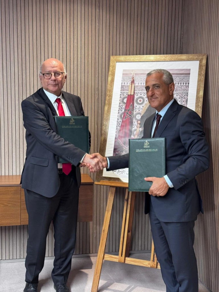 Two businessmen in suits shake hands after a formal agreement, each holding a green folder in a modern office setting.”] } {