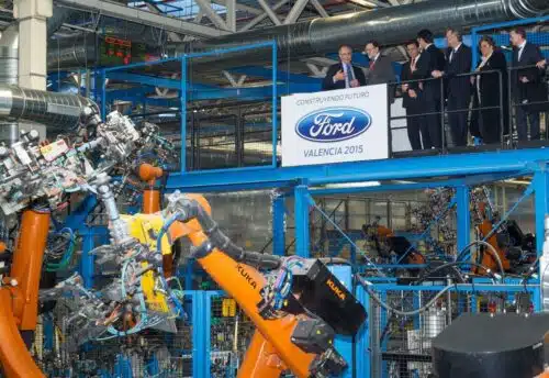 Ford Valencia 2015 event: executives on a viewing balcony overlook a robotic assembly line with orange robotic arms in a car production plant.