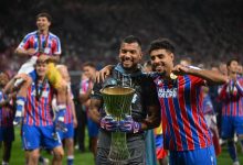 Two soccer players celebrate a win, smiling and holding a tall silver trophy on the field with teammates behind them in red-and-blue striped kits.