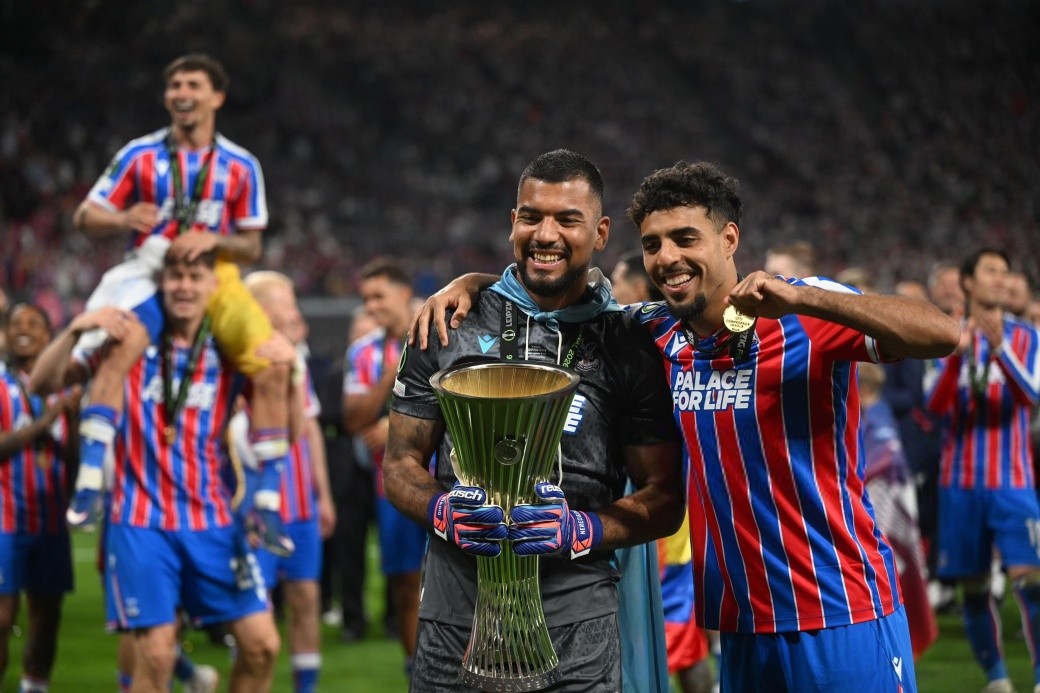 Two soccer players celebrate a win, smiling and holding a tall silver trophy on the field with teammates behind them in red-and-blue striped kits.