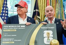 74144152_605 | MCG24 | موقع إلكتروني مغربي شامل متجدد على مدار الساعة Two men in suits hold a large mock FIFA World Cup 2026 ticket, with flags and a trophy nearby in a formal room. One wears a red cap partially visible in the frame.