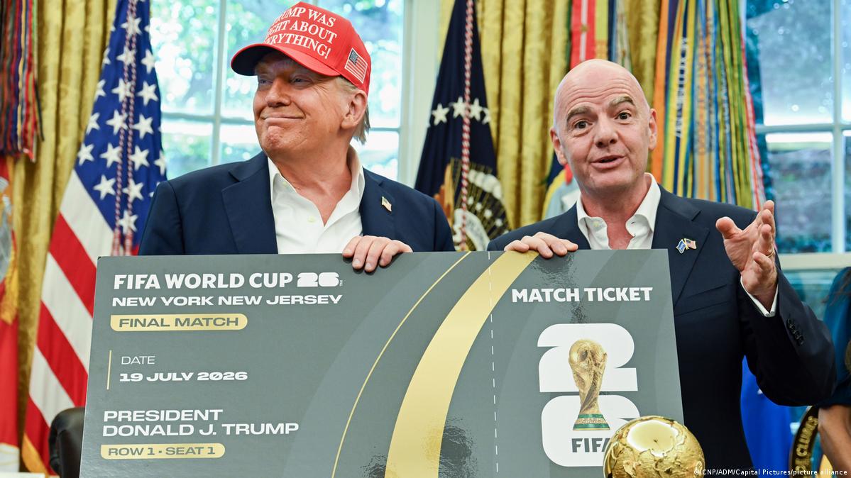 74144152_605 | MCG24 | موقع إلكتروني مغربي شامل متجدد على مدار الساعة Two men in suits hold a large mock FIFA World Cup 2026 ticket, with flags and a trophy nearby in a formal room. One wears a red cap partially visible in the frame.