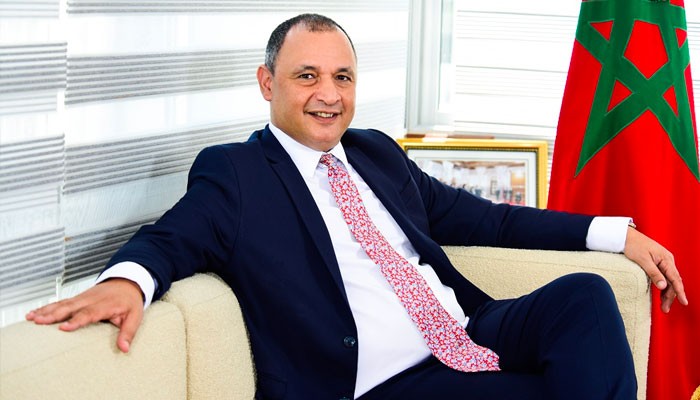 Man in a navy suit and red patterned tie sits on a beige sofa in an office, with a Moroccan flag in the background.