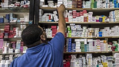 A person in a blue shirt reaches up to grab a box from high pharmacy shelves full of medicine boxes.